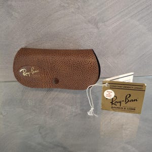 May include: A brown, textured Ray-Ban sunglasses case with a snap closure. The Ray-Ban logo is in gold. A tag with the Ray-Ban logo and text "CONSTANT DENSITY LENSES" is attached.