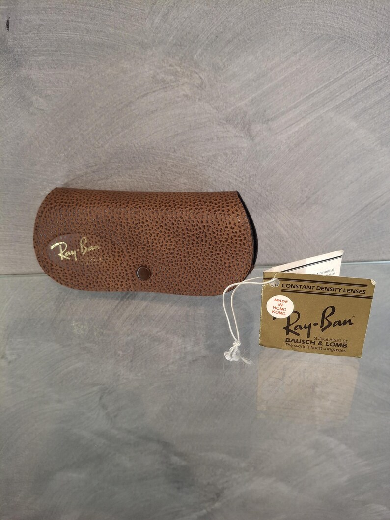 May include: A brown, textured Ray-Ban sunglasses case with a snap closure. The Ray-Ban logo is in gold. A tag with the Ray-Ban logo and text "CONSTANT DENSITY LENSES" is attached.