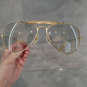 May include: Gold aviator sunglasses with clear lenses. The sunglasses are held in a hand.