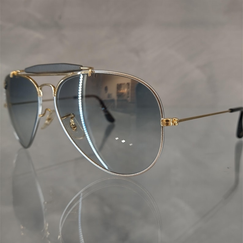 Puede incluir: Gafas de sol aviador doradas con lentes de degradado gris y una barra de cejas negra.