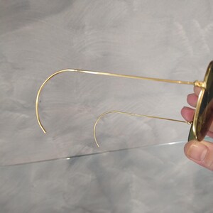 May include: Gold-tone round sunglasses with green lenses. The sunglasses are folded in half and the arms are bent into a curved shape.