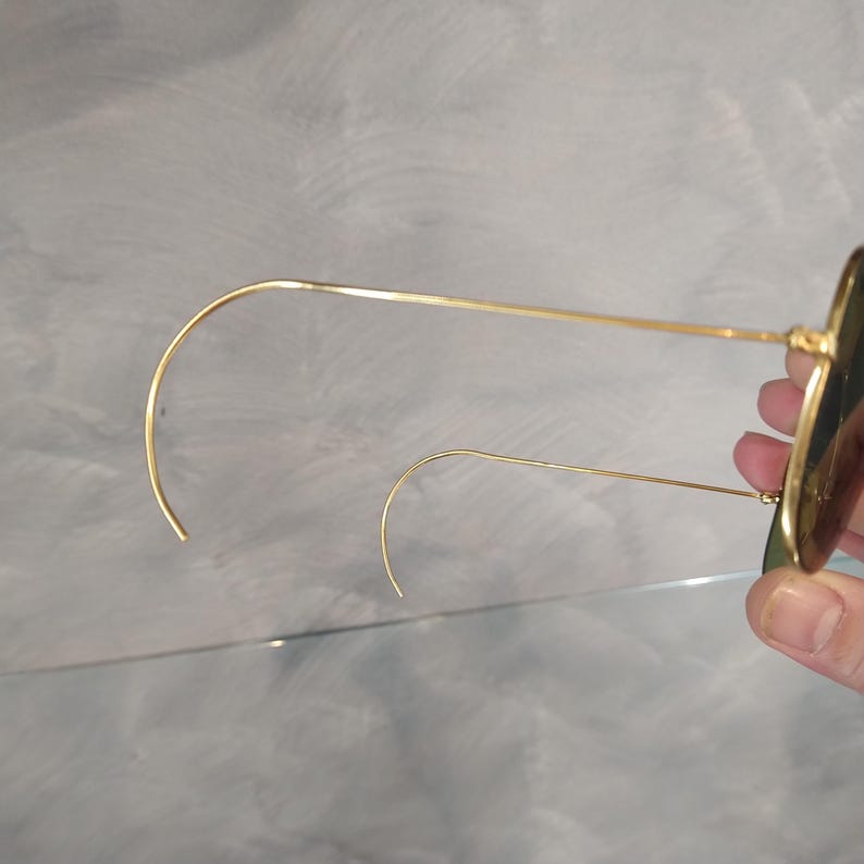 May include: Gold-tone round sunglasses with green lenses. The sunglasses are folded in half and the arms are bent into a curved shape.