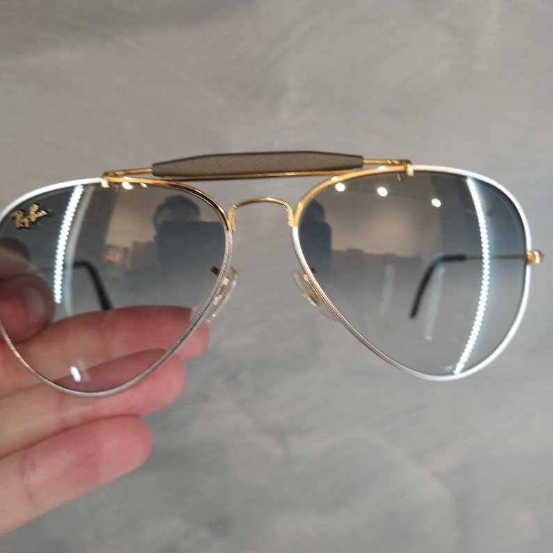 Puede incluir: Gafas de sol aviador con montura dorada, lentes de degradado gris y puente de cuero marr&oacute;n.