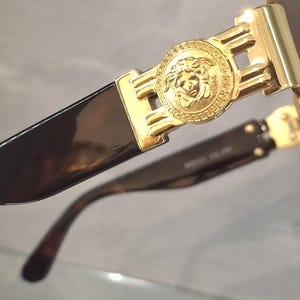 K&ouml;nnte beinhalten: Nahaufnahme einer goldfarbenen Sonnenbrille mit einem Medusenhaupt-Emblem auf dem Rahmen. Die Gl&auml;ser sind dunkel und die B&uuml;gel haben ein Schildpattmuster. Ein luxuri&ouml;ses Modeaccessoire.