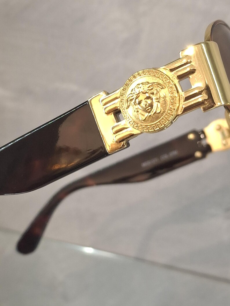 K&ouml;nnte beinhalten: Nahaufnahme einer goldfarbenen Sonnenbrille mit einem Medusenhaupt-Emblem auf dem Rahmen. Die Gl&auml;ser sind dunkel und die B&uuml;gel haben ein Schildpattmuster. Ein luxuri&ouml;ses Modeaccessoire.