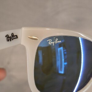 Pu&ograve; includere: Occhiali da sole bianchi con lenti blu. Il logo Ray-Ban &egrave; visibile sulla lente e sull'asta. La montatura &egrave; di colore bianco brillante e le lenti riflettono l'ambiente.