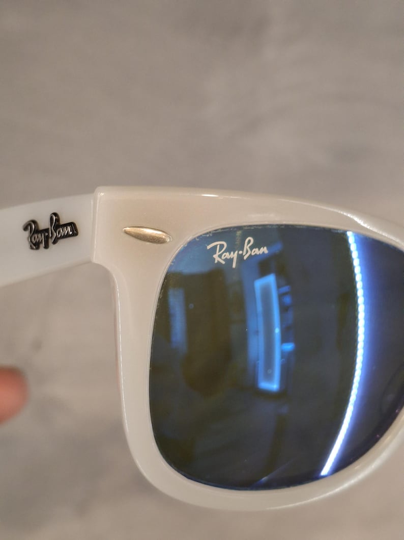 Pu&ograve; includere: Occhiali da sole bianchi con lenti blu. Il logo Ray-Ban &egrave; visibile sulla lente e sull'asta. La montatura &egrave; di colore bianco brillante e le lenti riflettono l'ambiente.
