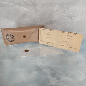 May include: A brown leather Ray-Ban glasses case with a snap closure. The case is open and shows the interior lining. There are two white paper tags with the text "BAUSCH & LOMB" and "Ray-Ban" on them. The tags contain the text "(LEFT)" and "(RIGHT)" with the catalogue number "631 47 02".