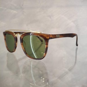 May include: Brown tortoiseshell sunglasses with green lenses. The sunglasses have a gold-colored metal brow bar and temples. The frames are a classic shape, suitable for various face shapes. The lenses are a dark green color.