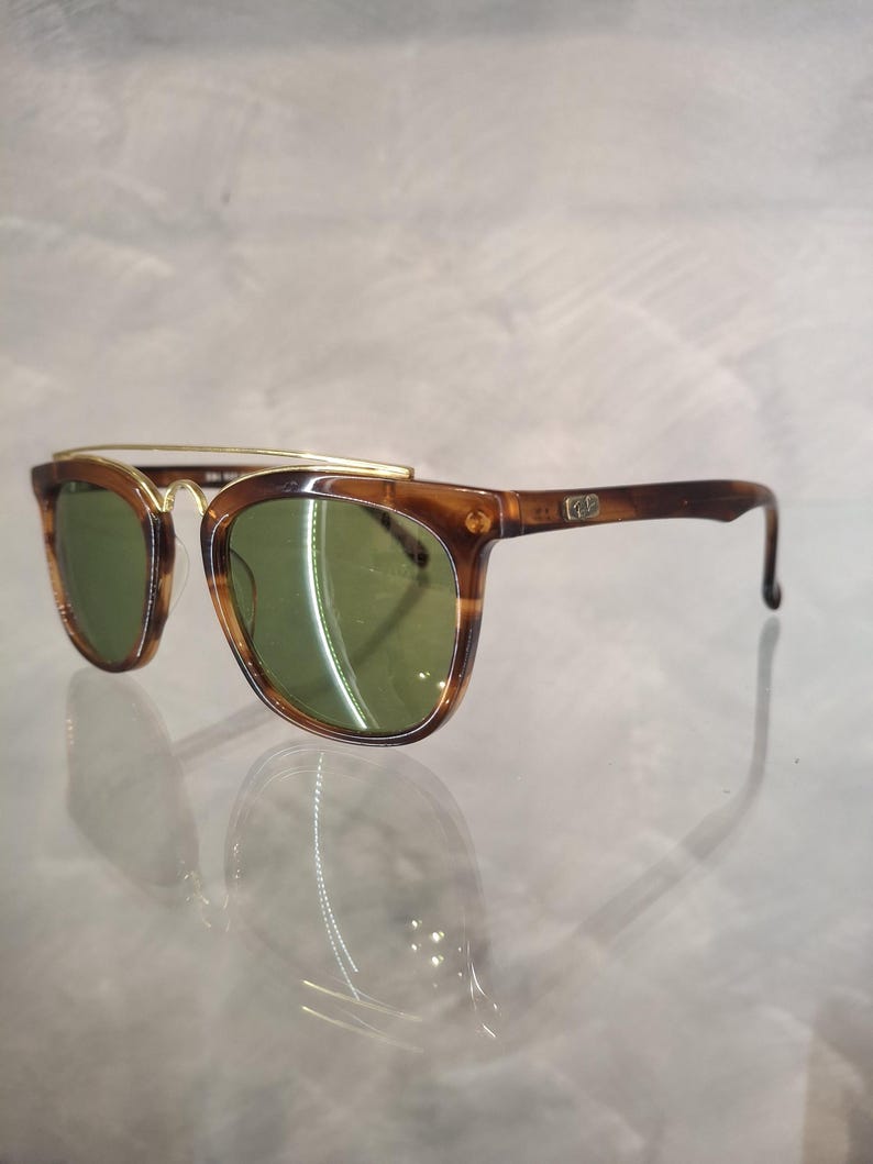 May include: Brown tortoiseshell sunglasses with green lenses. The sunglasses have a gold-colored metal brow bar and temples. The frames are a classic shape, suitable for various face shapes. The lenses are a dark green color.