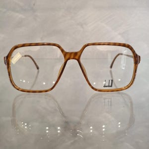 May include: Brown tortoiseshell eyeglasses with a square frame. The glasses have a clear lens and thin metal arms. The brand name "dunhill" is visible on the right lens. A price tag is attached to the left side of the frame.
