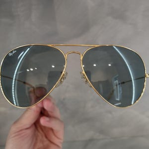 Puede incluir: Gafas de sol aviador doradas con lentes grises oscuros. Las gafas tienen el logotipo de Ray-Ban en la lente izquierda.