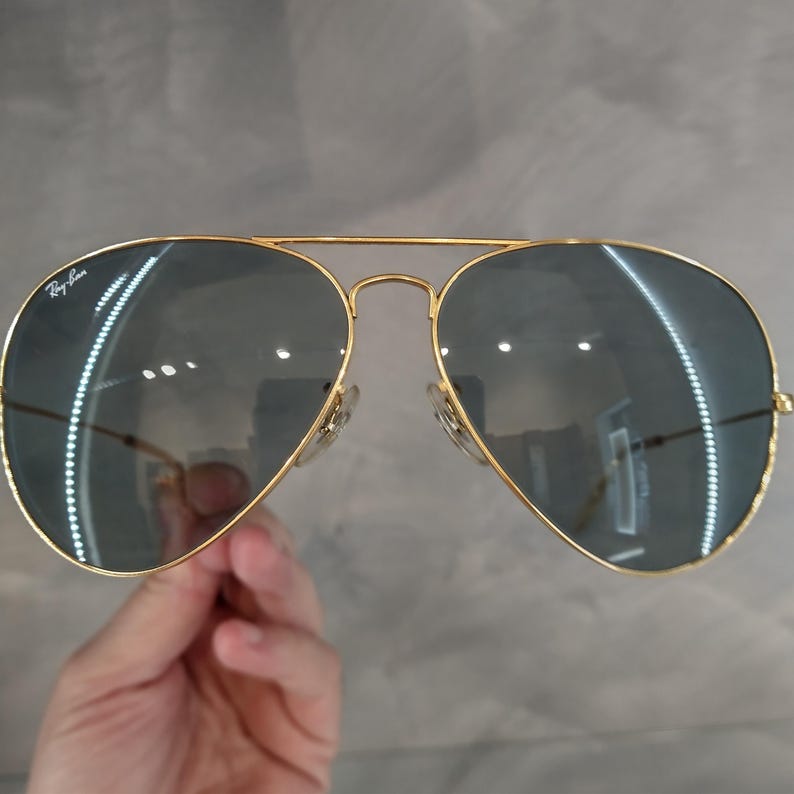 Puede incluir: Gafas de sol aviador doradas con lentes grises oscuros. Las gafas tienen el logotipo de Ray-Ban en la lente izquierda.