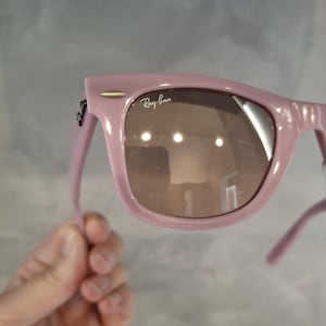 Puede incluir: Gafas de sol rosas con lentes marrones. El logotipo de Ray-Ban es visible en la lente. Las gafas de sol est&aacute;n sujetas por una mano.