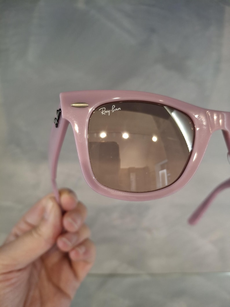 Puede incluir: Gafas de sol rosas con lentes marrones. El logotipo de Ray-Ban es visible en la lente. Las gafas de sol est&aacute;n sujetas por una mano.