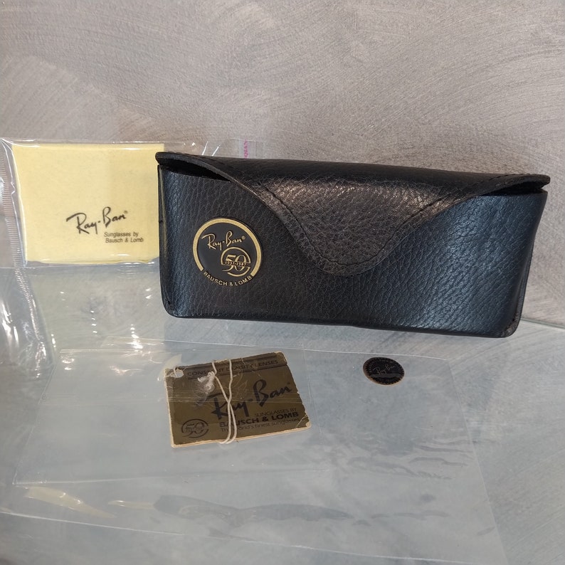 May include: A black leather Ray-Ban sunglasses case with a gold Ray-Ban logo and the text "50th Anniversary" and "Bausch & Lomb".