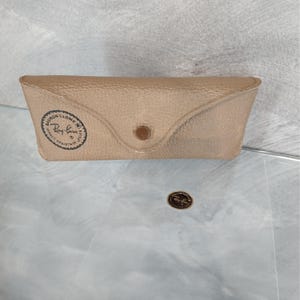 May include: A tan Ray-Ban glasses case with a snap closure. The case has a textured surface and a circular logo on the front. A small Ray-Ban logo sticker is also visible.