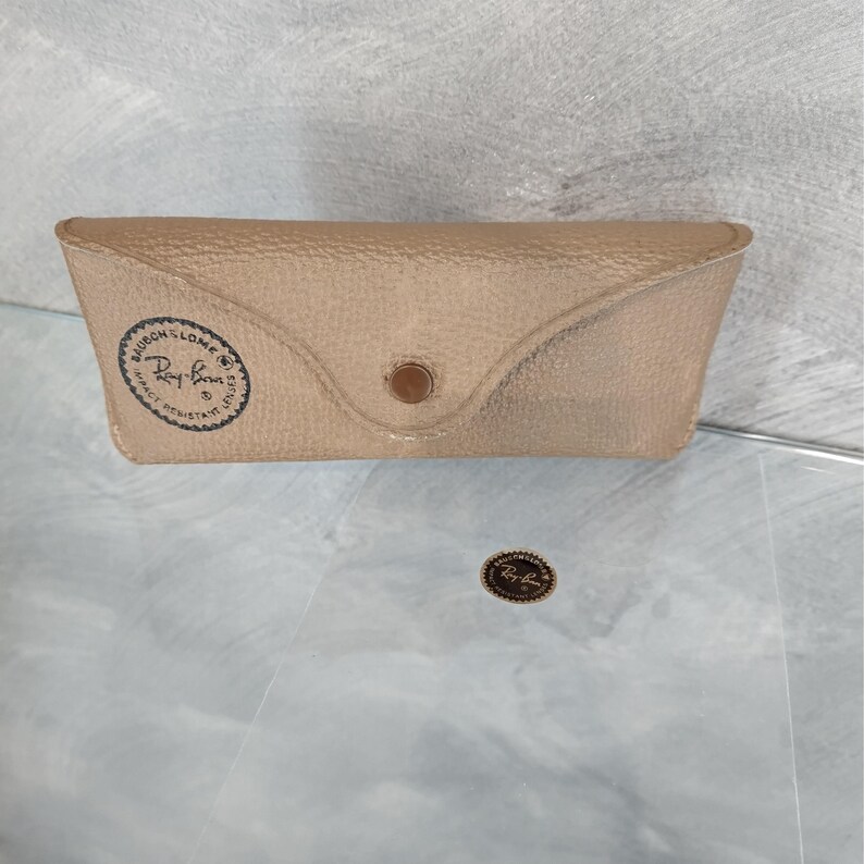 May include: A tan Ray-Ban glasses case with a snap closure. The case has a textured surface and a circular logo on the front. A small Ray-Ban logo sticker is also visible.