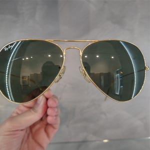 Puede incluir: Gafas de sol aviador doradas con lentes verdes. El logotipo de Ray-Ban es visible en la lente izquierda.