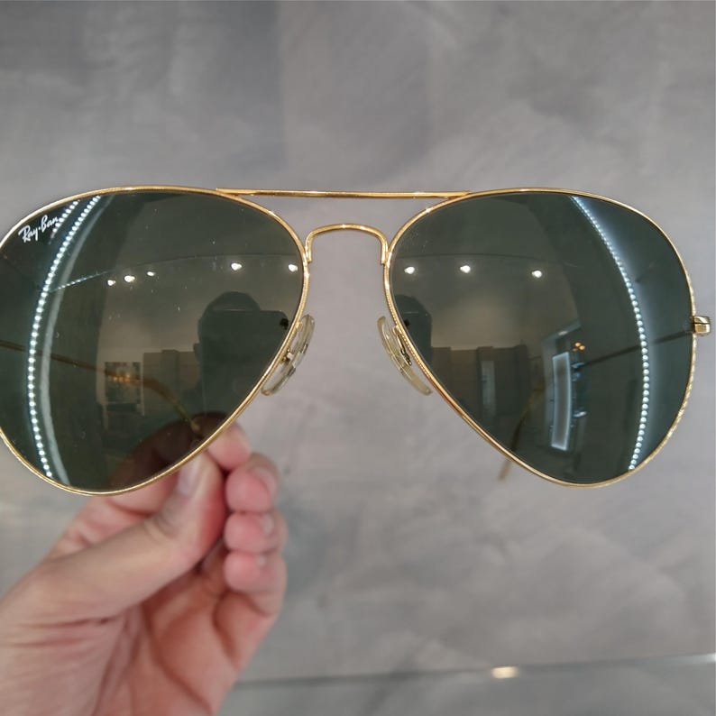 Puede incluir: Gafas de sol aviador doradas con lentes verdes. El logotipo de Ray-Ban es visible en la lente izquierda.