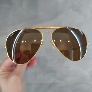 May include: Gold aviator sunglasses with brown lenses. The sunglasses have a Ray-Ban logo on the left lens.