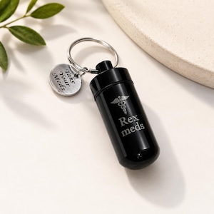Op de afbeelding: Een zwarte pillenhouder met een zilveren sleutelhanger en een ronde tag met de tekst "Take Your Meds". De pillenhouder heeft de tekst "Rex meds" en een medisch symbool. De houder is cilindrisch.