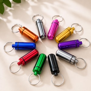 Op de afbeelding: Een verzameling kleurrijke, cilindervormige sleutelhangers in diverse metallic afwerkingen, waaronder oranje, zilver, roze, goud, paars, blauw, rood, groen en zwart. Elke sleutelhanger heeft een schroefdop en een zilveren sleutelring.
