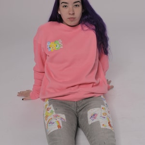 May include: A pink sweatshirt with a cartoon character patch on the front, paired with gray jeans with colorful patches. The jeans have distressed details.
