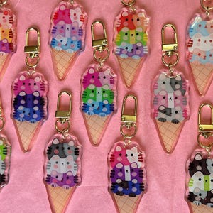 May include: A selection of colourful acrylic keyrings shaped like ice cream cones, each topped with a stack of cat faces. The keyrings feature gold-coloured clasps and are arranged on a pink surface.