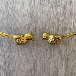 May include: Two gold bird-shaped drawer pulls, facing each other, with detailed wings and beaks.