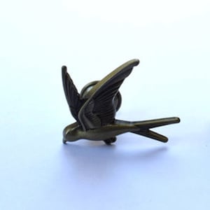 May include: Bronze-toned bird brooch with detailed wing and feather design. The bird is depicted in flight, with a pointed beak and a long, slender tail. The brooch is set against a plain white background.