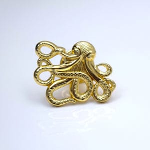 May include: A gold-coloured octopus pin with a detailed textured design. The octopus has eight tentacles and is curled up in a compact shape.