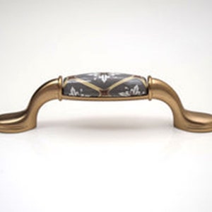 May include: A cabinet pull with a gold finish and a decorative centre. The centre section is grey with a white floral pattern and gold accents. The pull has a curved design, suitable for furniture.