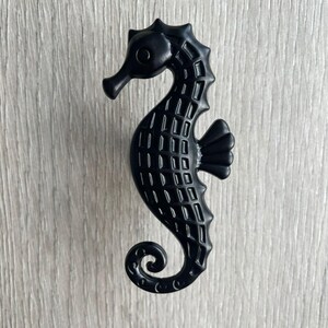 May include: Black metal seahorse door knob or cabinet pull. The seahorse is detailed with a textured pattern and has a curled tail.