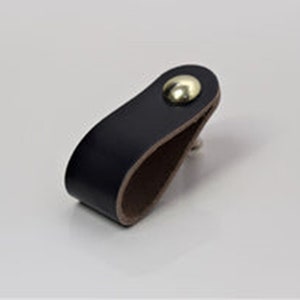 May include: A black leather drawer pull with a brass knob. The pull is shaped like a loop and has a brown leather backing.