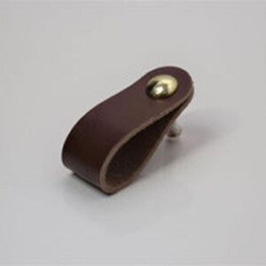 May include: A brown leather cabinet pull with a brass knob. The pull is shaped like a loop and is designed to be mounted on a cabinet door.