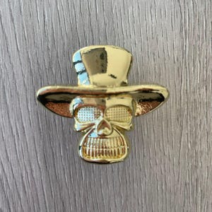 May include: A gold-coloured skull wearing a top hat. The skull has a detailed design with a wide grin and eye sockets.