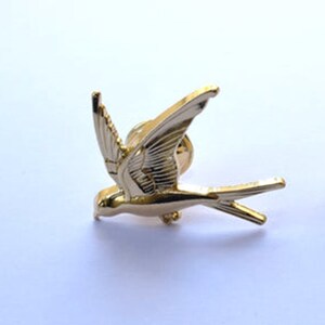 May include: A gold-coloured metal bird pin in the shape of a swallow. The pin is in flight with its wings spread out.