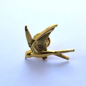 May include: A gold-coloured metal bird pin with outstretched wings, featuring detailed feather patterns. The pin is designed in a flying pose, suggesting movement. A decorative accessory.
