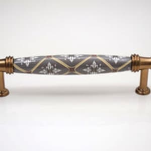 May include: A grey ceramic cabinet pull with a gold finish and a white and brown geometric pattern. The pull is approximately 12.7 cm long.