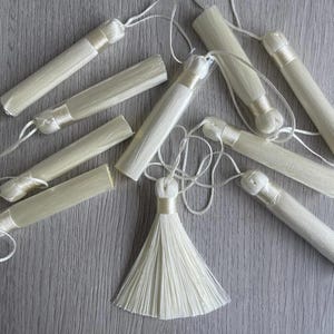 May include: A collection of ten white tassel trims with a single larger white tassel in the centre. The tassels are made of soft, fluffy fibres and are attached to thin white cords.