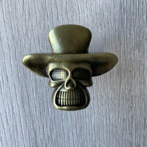 May include: A gold-toned metal skull wearing a top hat. The skull has a detailed face with a toothy grin.