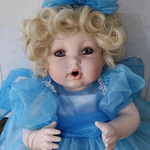 May include: A porcelain doll with blonde curly hair and blue eyes. The doll wears a light blue dress with a matching bow. The doll's arms are outstretched, and it has a surprised expression. The doll is part of the "Miracle Children" collection.
