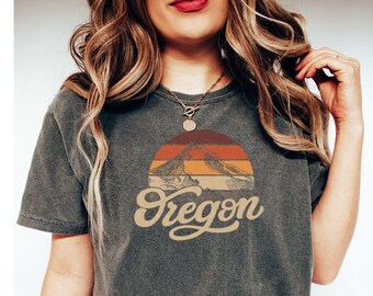 May include: A dark grey cropped t-shirt featuring a vintage-style graphic of mountains and a sunset. The graphic includes the word "Oregon" in a stylised font.