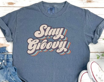 Könnte beinhalten: Ein blaues T-Shirt mit dem Text "Stay Groovy" in einer Retro-Schriftart mit mehrfarbigen Umrissen. Das Shirt liegt auf einer weißen Holzoberfläche mit einem Paar weißen Canvas-Schuhen und einer blauen Jeans.