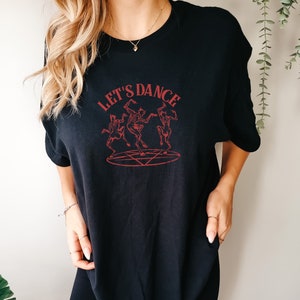 Op de afbeelding: Zwart T-shirt met een rode afbeelding van skeletten die rond een pentagram dansen. De tekst "LET'S DANCE" staat boven de afbeelding.