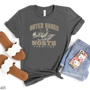 Pode incluir: Uma camiseta cinza escura com o texto "Outer Banks North Carolina" em uma fonte de estilo vintage. A camiseta apresenta um gr&aacute;fico de um farol e uma cena de praia.