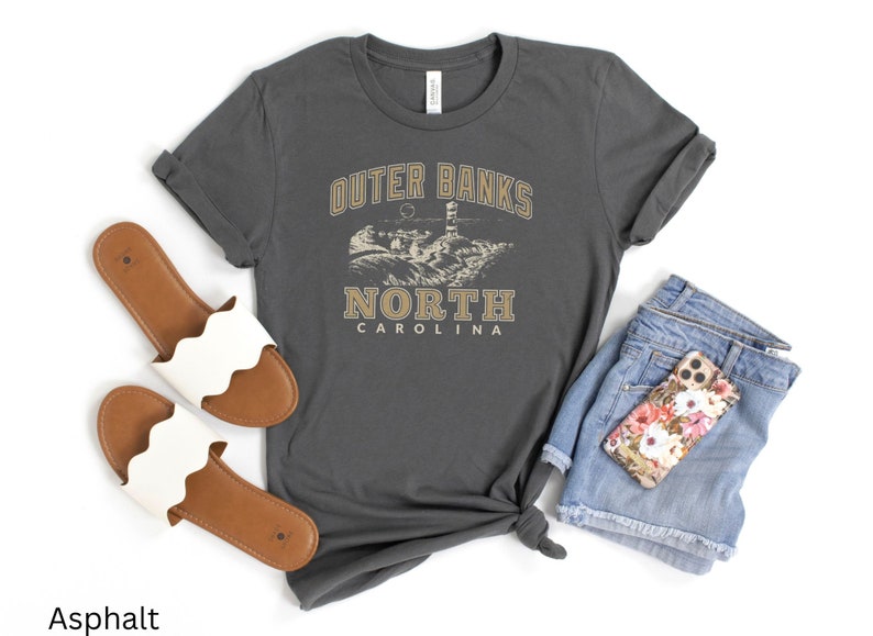Pode incluir: Uma camiseta cinza escura com o texto "Outer Banks North Carolina" em uma fonte de estilo vintage. A camiseta apresenta um gr&aacute;fico de um farol e uma cena de praia.
