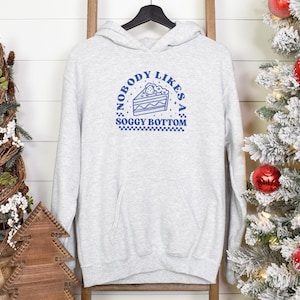 May include: Light gray hoodie with a front pocket and a blue graphic that says "NOBODY LIKES A SOGGY BOTTOM" with a cake slice illustration. The hoodie is hanging on a wooden ladder with Christmas decorations in the background.