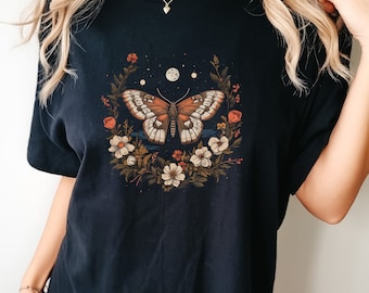 May include: Black t-shirt with a floral wreath design featuring a butterfly and moons. The butterfly has brown, white, and orange wings.